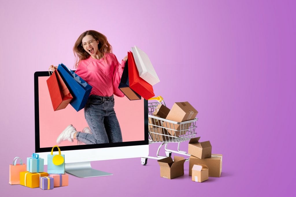 Bright young woman in pink sweatshirt smiling and holding shopping bags coming out of a computer monitor, surrounded by shopping boxes and a toy shopping cart against a purple background.
